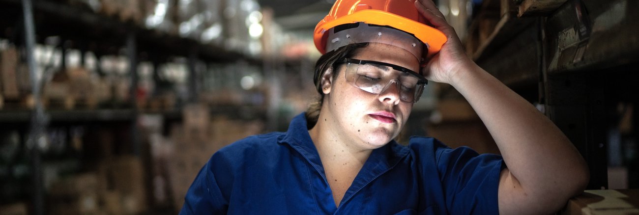 Arbeiterin mit Helm, Schutzbrille und Tablet in einem Logistikzentrum mit Regalen hat einen Arm aufgestützt und die Augen geschlossen © iStock/FG Trade