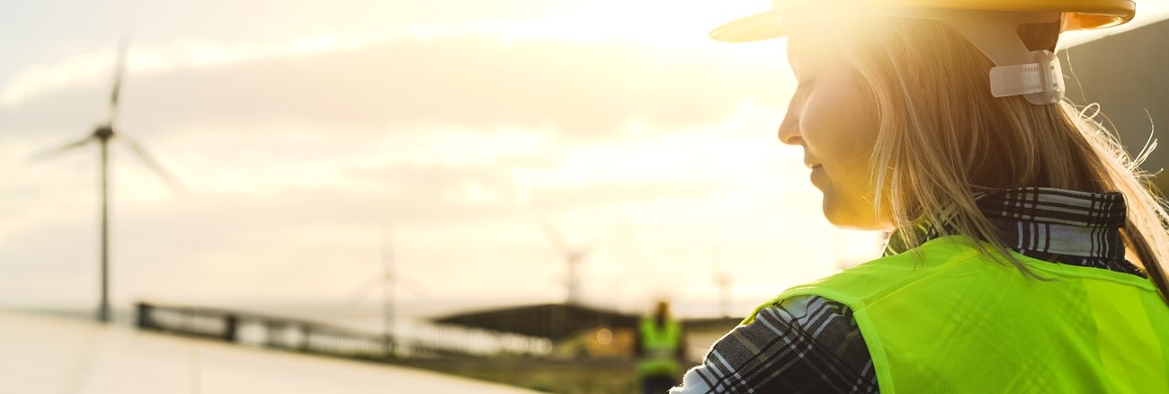 Arbeiterin mit Helm an einer Photovoltaik-Anlage, dahinter Windkraftturbinen © envato/biasciolialessandro Arbeiterin mit Helm an einer Photovoltaik-Anlage, dahinter Windkraftturbinen © envato/biasciolialessandro