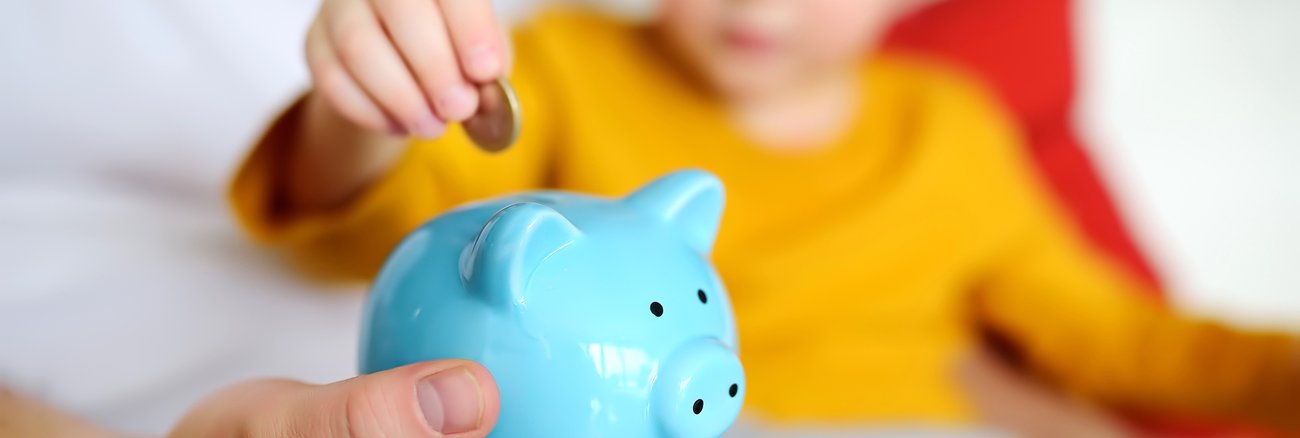 Junge mit gelbem Pullover steckt Euro-Münze in ein hellblaues Sparschwein © iStock/Finanzwissen Junge mit gelbem Pullover steckt Euro-Münze in ein hellblaues Sparschwein © iStock/Finanzwissen