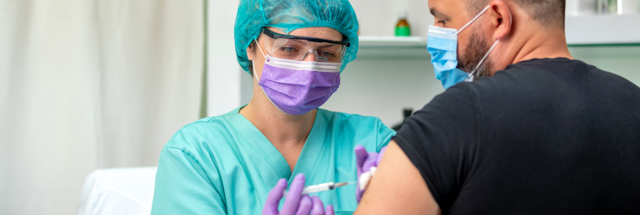 Krankenschwester mit Schutzbrille und Maske verabreicht einem Mann mit Maske eine Injektion @ iStock/zoranm Krankenschwester mit Schutzbrille und Maske verabreicht einem Mann mit Maske eine Injektion @ iStock/zoranm