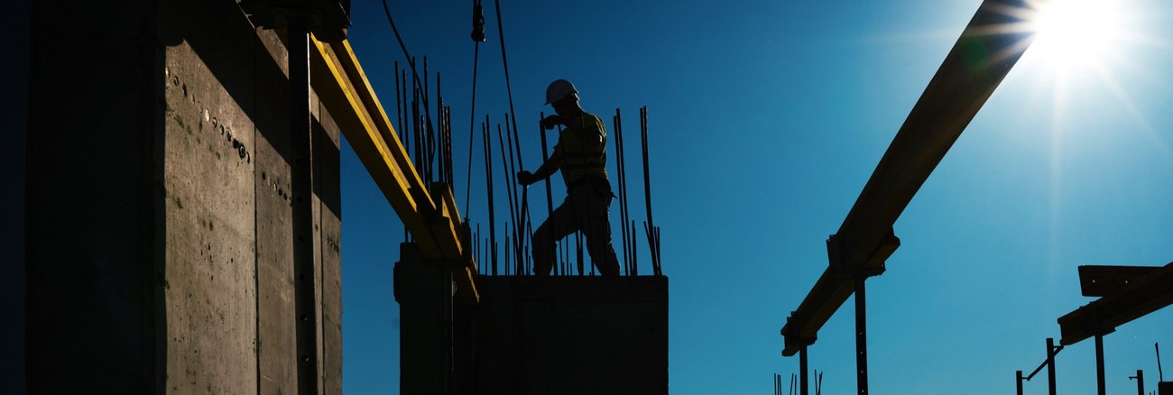 Silhouette eines Bauarbeiters auf einer Baustelle mit Stahlträgern, Kran und Betonpfeilern © iStock/gilaxia Silhouette eines Bauarbeiters auf einer Baustelle mit Stahlträgern, Kran und Betonpfeilern © iStock/gilaxia