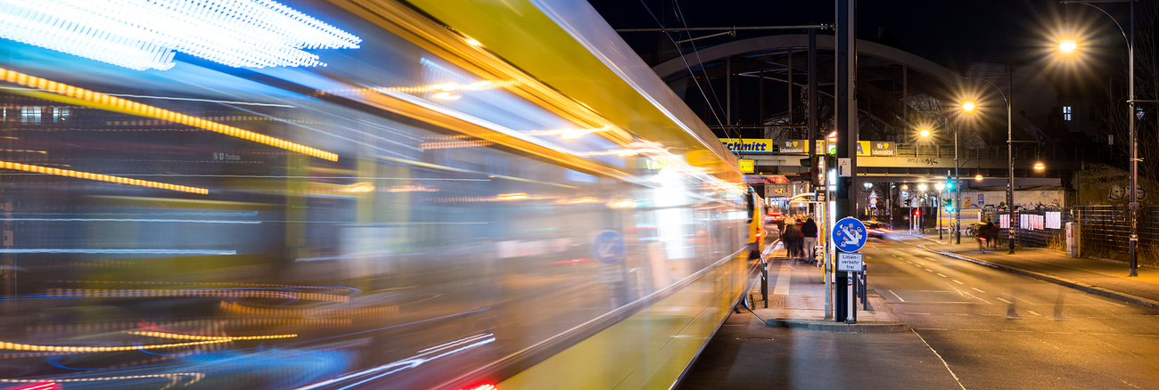 Eine gelbe Straßenbahn fährt nachts in eine Haltestelle ein, die von Straßenlaternen erleuchtet wird. Im Hintergrund ist eine Bahnbrücke zu sehen. Foto: © istock.com/holgs