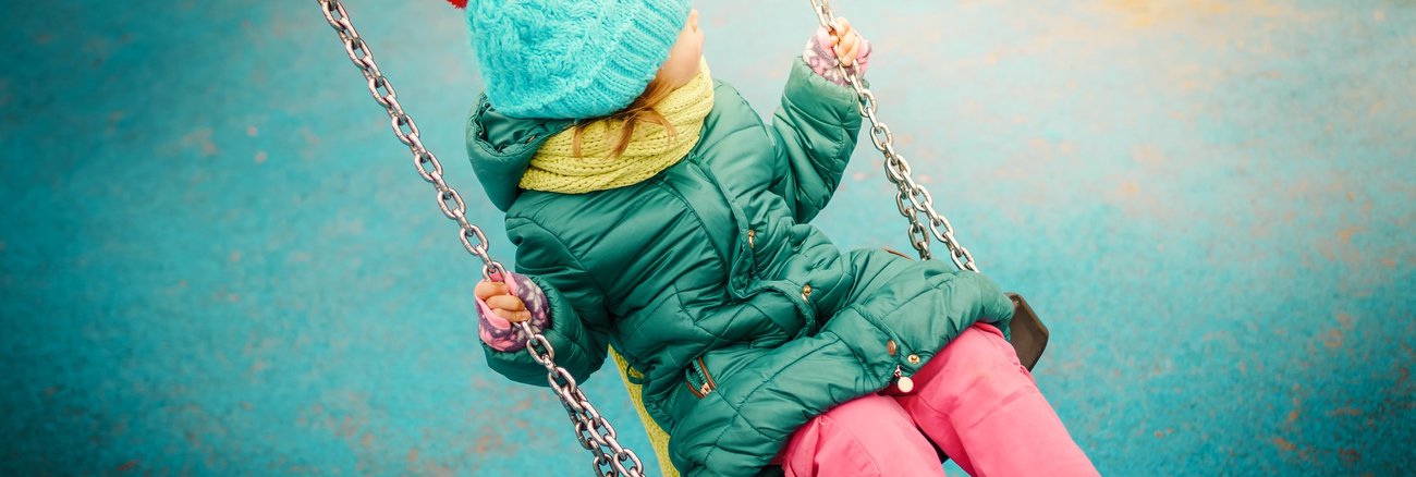 Ein Mädchen bei Regen mit Pudelmütze und Gummistiefeln auf einer Schaukel © iStock/oska25