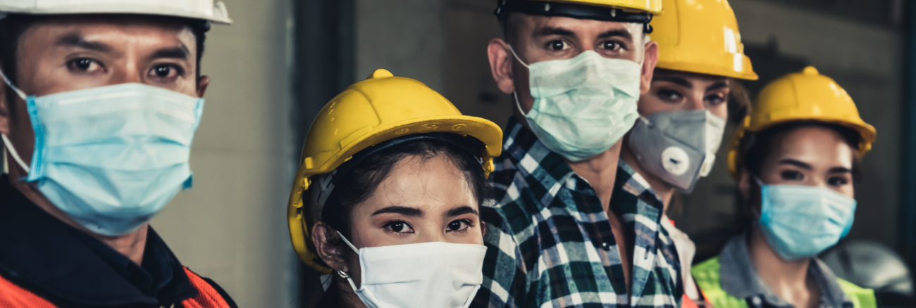 Arbeiter mit Helm und Mund-Nase-Schutzmaske stehen in einer Reihe und blicken in die Kamera. Foto: © istock.com / Blue Planet Studio Arbeiter mit Helm und Mund-Nase-Schutzmaske stehen in einer Reihe und blicken in die Kamera. Foto: © istock.com / Blue Planet Studio