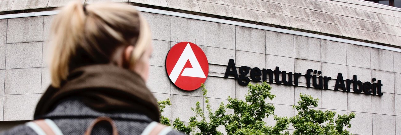 Frau mit Rucksack vor einem Gebäude mit dem Logo und der Aufschrift Agentur für Arbeit © picture alliance/imageBROKER|Jan Tepass