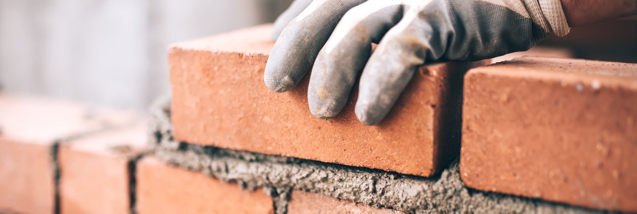 Eine Hand mit Handschuh hält einen Ziegelstein auf einer unvollständigen Wand mit frischem Mörtel © iStock/Bogdanhoda