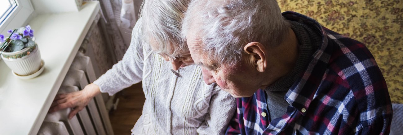 Eine ältere Frau hält eine Rechnung und fasst auf einen Heizkörper, daneben ein älterer Mann mit einer Decke auf den Beinen © iStock/Marina113