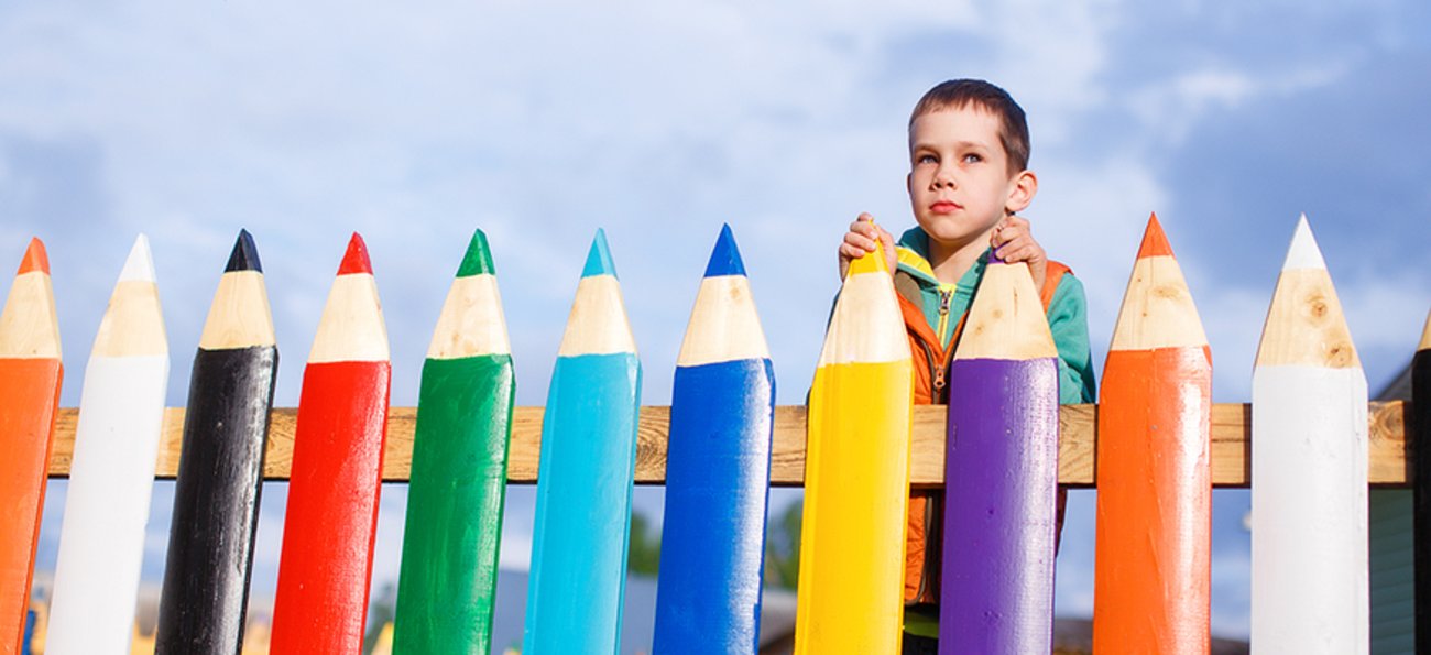 Kind blickt nachdenklich über einen Zaun, dessen Latten wie Buntstifte aussehen. Foto: © istock.com/EvgeniiAnd