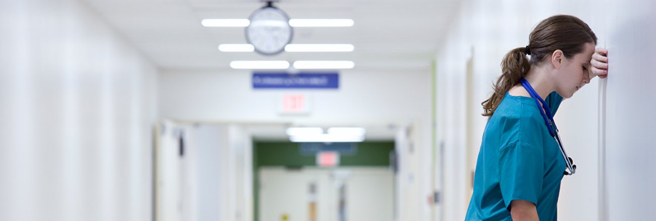 Eine erschöpfte Pflegekraft lehnt an der Wand eines Flurs im Krankenhaus. Foto: © istock.com/FangXiaNuo