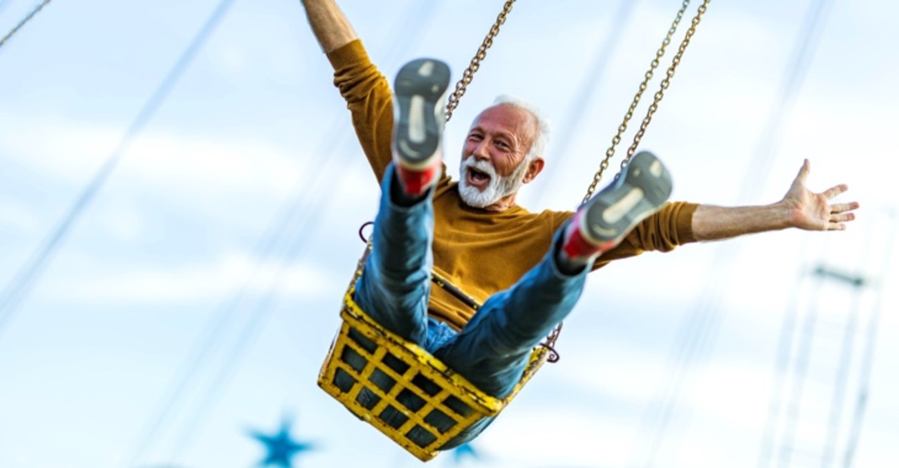 Älterer Mann mit Bart und ausgestreckten Armen sitzt lachend im Kettenkarussell © iStock/skynesher