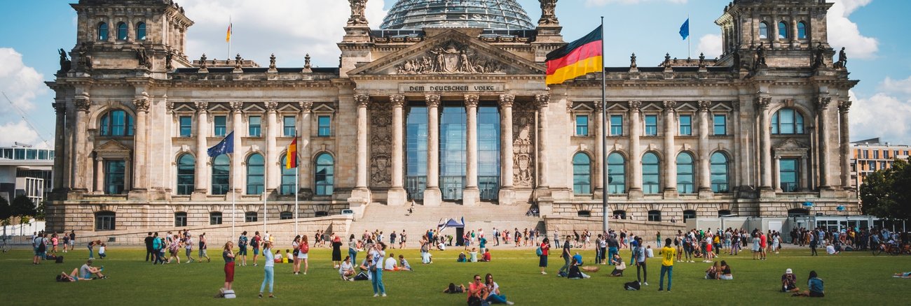 Menschen stehen und sitzen auf der Wiese vor dem Reichstagsgebäude in Berlin © iStock/hanohiki