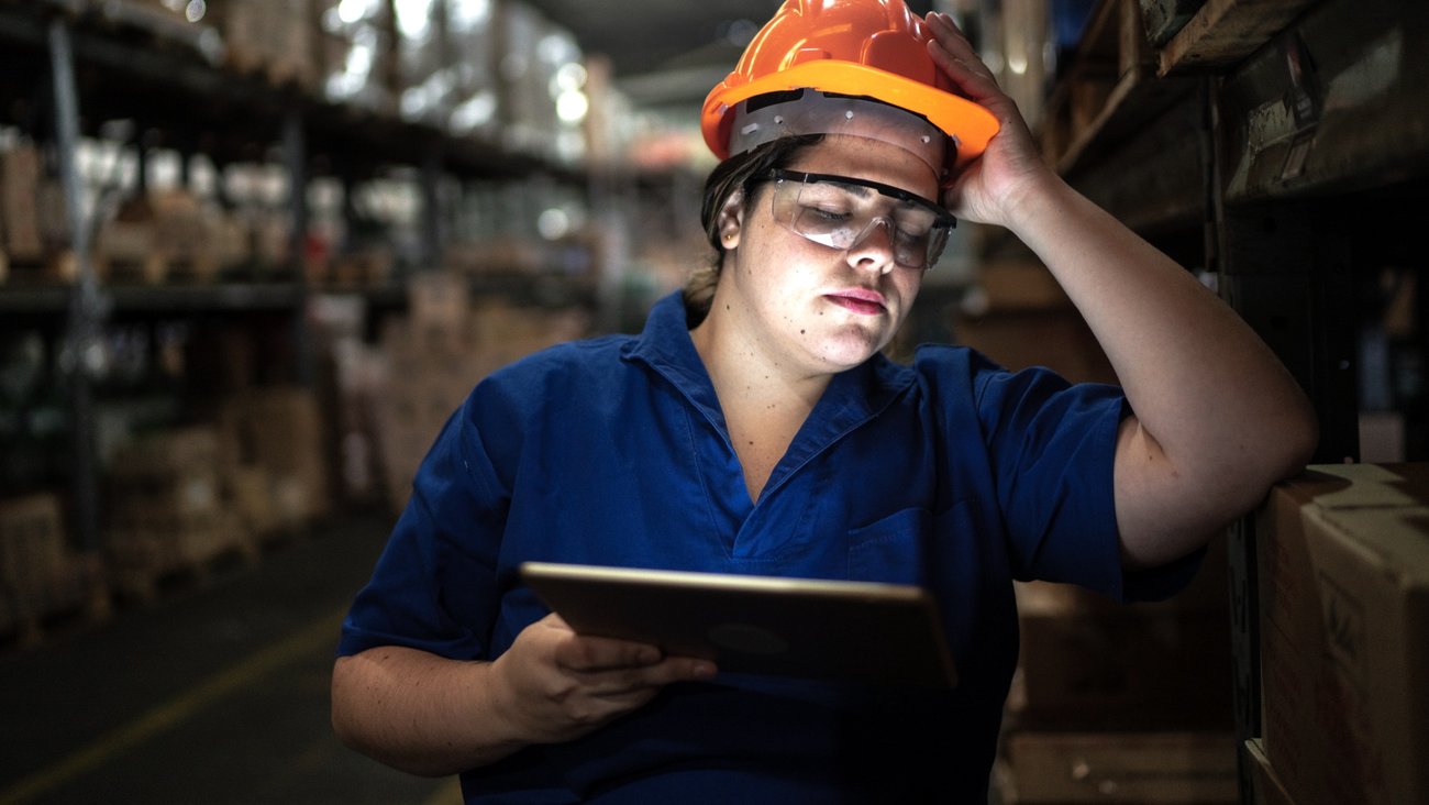 Arbeiterin mit Helm, Schutzbrille und Tablet in einem Logistikzentrum mit Regalen hat einen Arm aufgestützt und die Augen geschlossen © iStock/FG Trade
