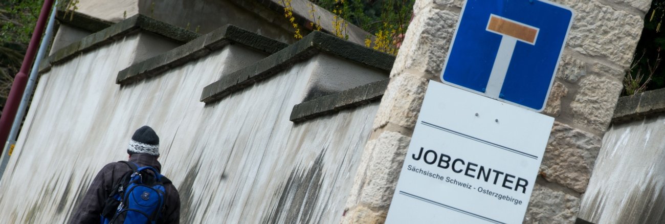 Ein Mann läuft an einem Sackgassen-Schild, das über einem Jobcenter-Schild angebracht ist, vorbei © Arno Burgi/dpa