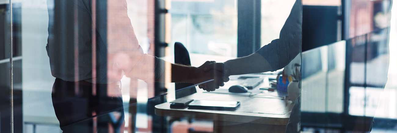 Zwei Männer in legerer Businesskleidung geben sich hinter der Glaswand eines Büros einen Handschlag. | Foto: © istock.com/gradyreese